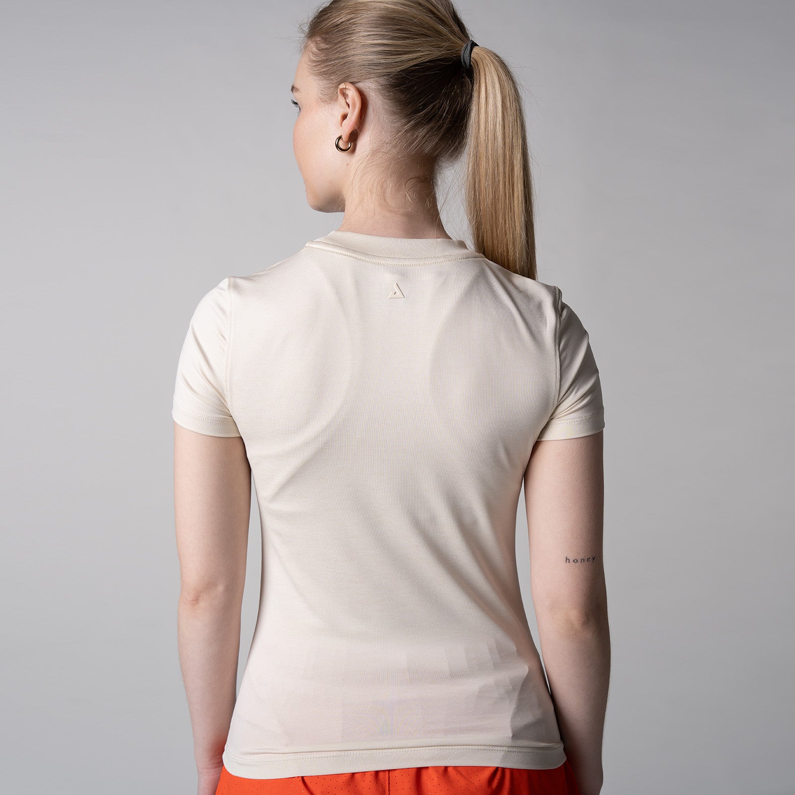 Gray background, image of the back of a female model wearing a JOOLA women's club short sleeve, sandstone color.