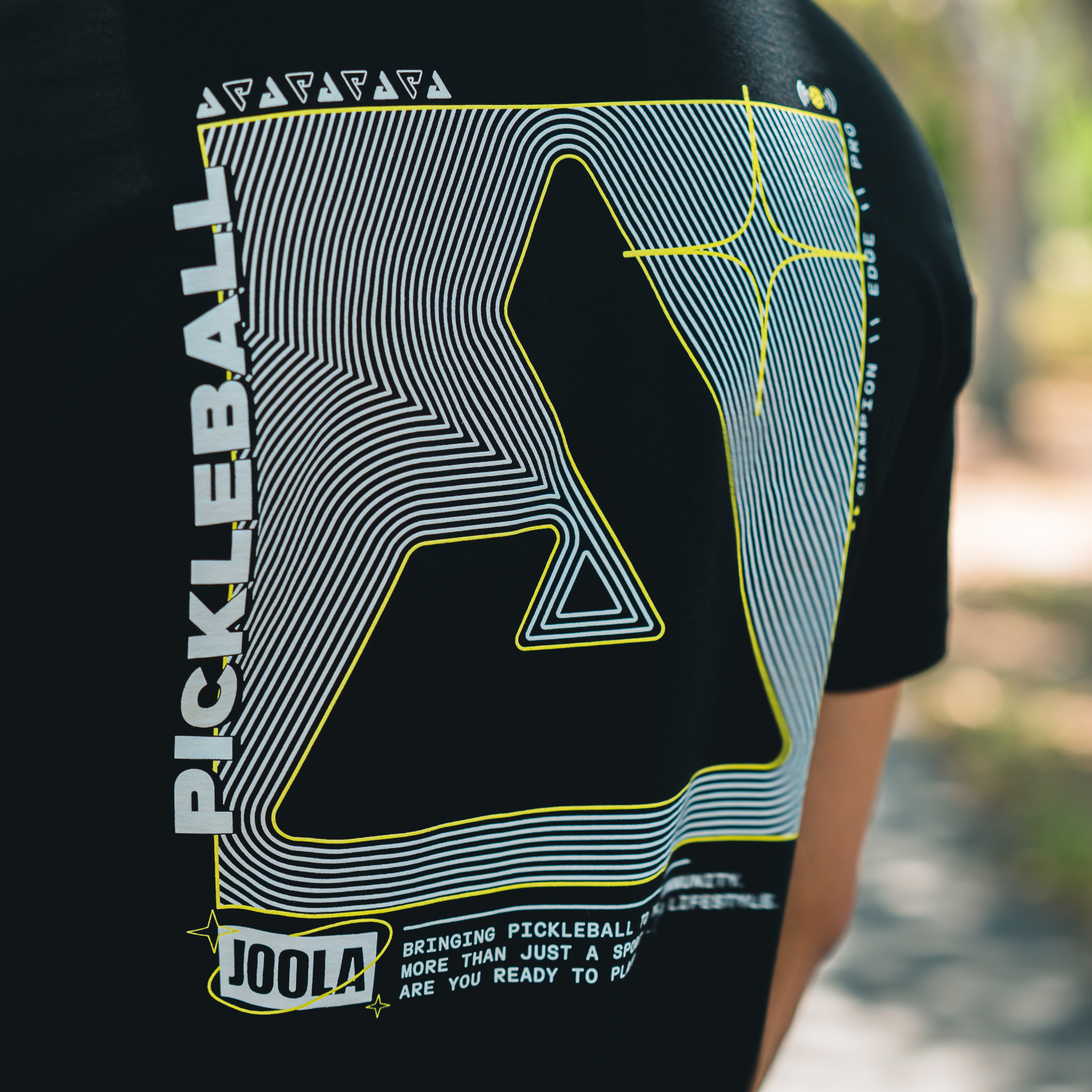 JOOLA Black t-shirt with pickleball design and text on a blurred outdoor background