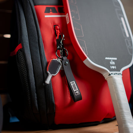 Agassi Vision red pickleball paddle bag with an Agassi Pro pickleball paddle and keychain on a wooden surface.