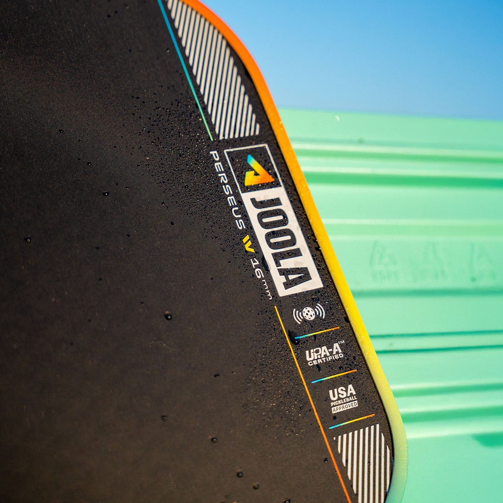 Close up image of the JOOLA Perseus Summer Daze Pickleball paddle with black surface, orange, blue, green and yellow color gradient on the rim, and white handle with a green and blue unfocused image in the background.