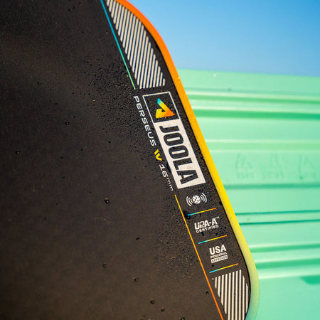 Close up image of the JOOLA Perseus Summer Daze Pickleball paddle with black surface, orange, blue, green and yellow color gradient on the rim, and white handle with a green and blue unfocused image in the background.