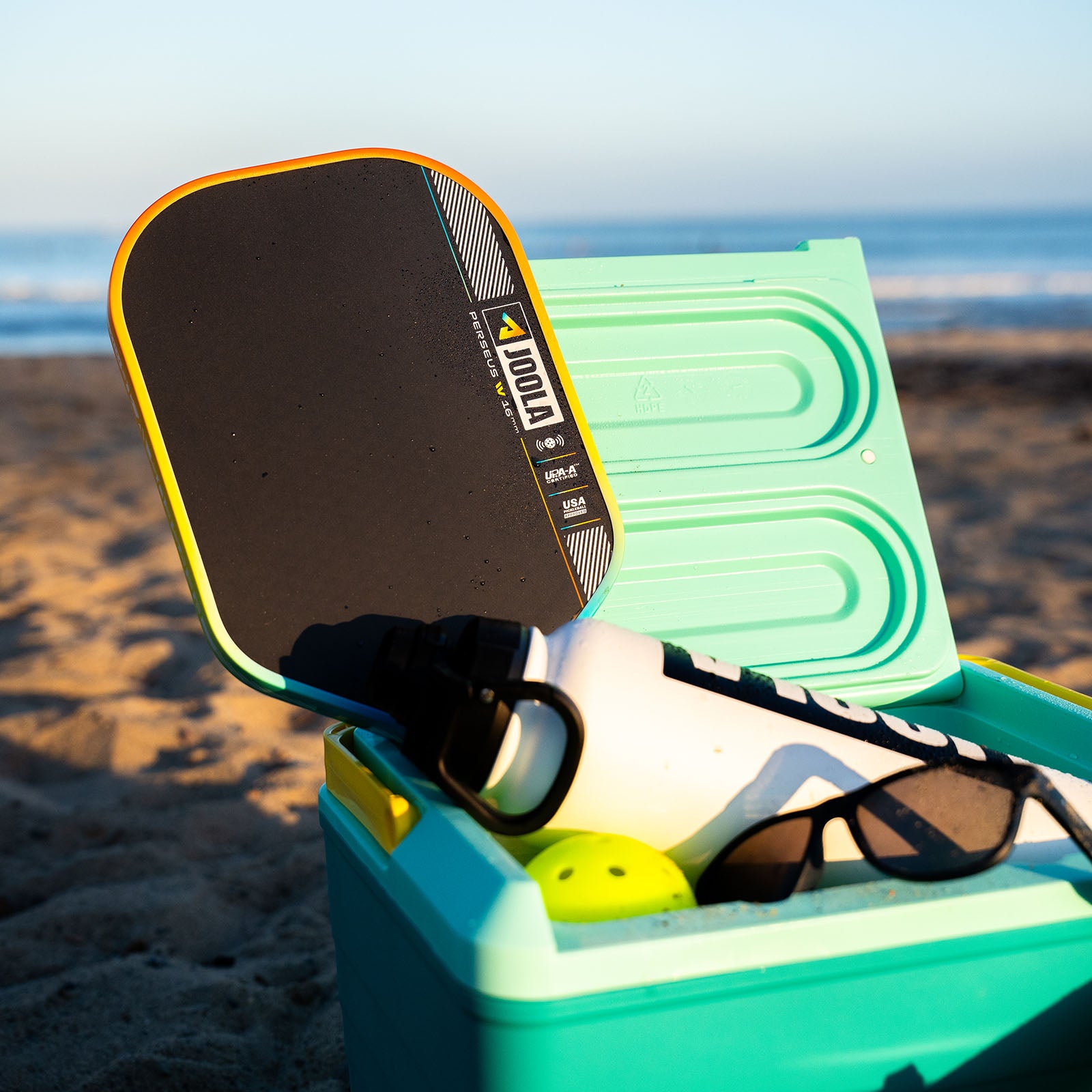 JOOLA Perseus Summer Daze Pickleball paddle with black surface, orange, blue, green and yellow color gradient on the rim, and white handle in an open cooler with items including sunglasses, and a bottle on a beach.