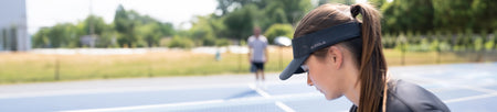 Horizontal banner image showing two JOOLA apparel models on a pickleball court. In the foreground is a female model wearing a JOOLA Visor.