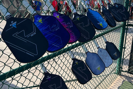 An array of JOOLA paddles displayed on the chain fence of the pickleball court