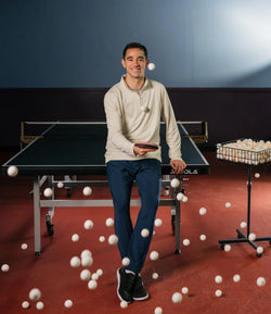 Image of Hugo Calderano hitting a ping pong ball in the air sitting on a table tennis table. Category