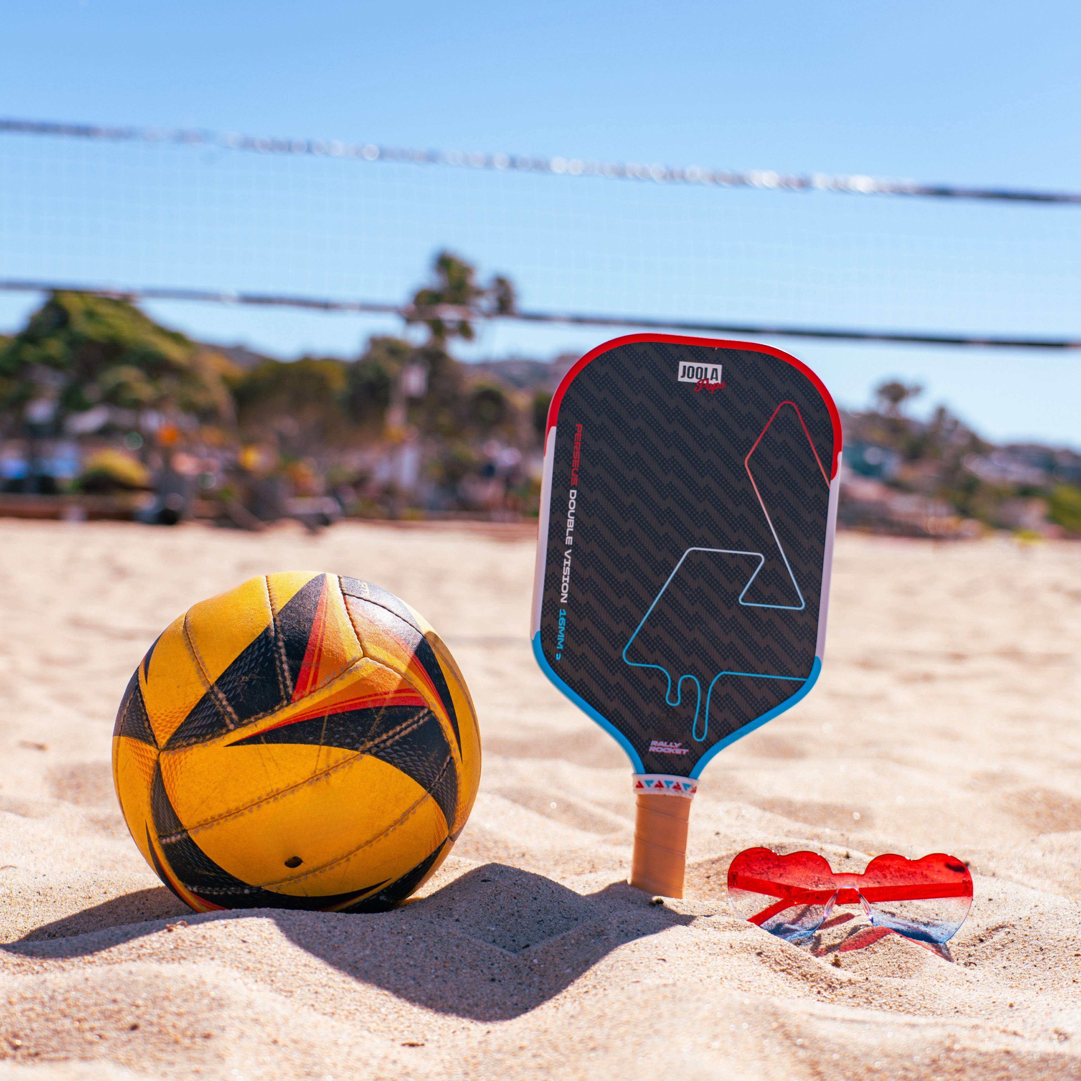 Beach volleyball ball and JOOLA Rally Rocket pickleball paddle on sand with volleyball net in background