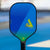 The JOOLA Wayfinder pickleball paddle with a blue and green gradient face, standing upright on a blue pickleball court with a blurred background.