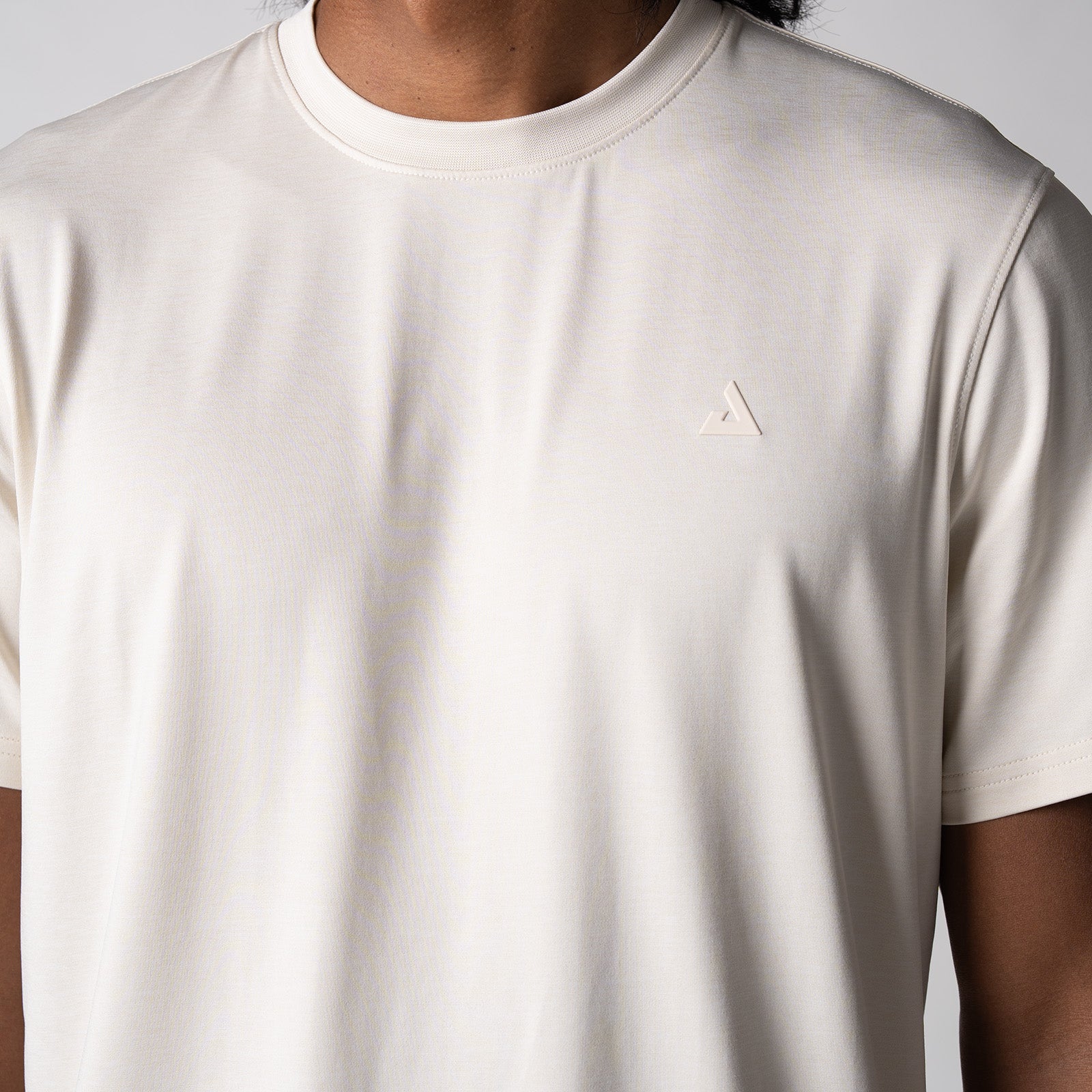Gray background, close up image of the JOOLA trinity logo over the left chest on a male model who is wearing the JOOLA men's club short sleeve, sandstone color.