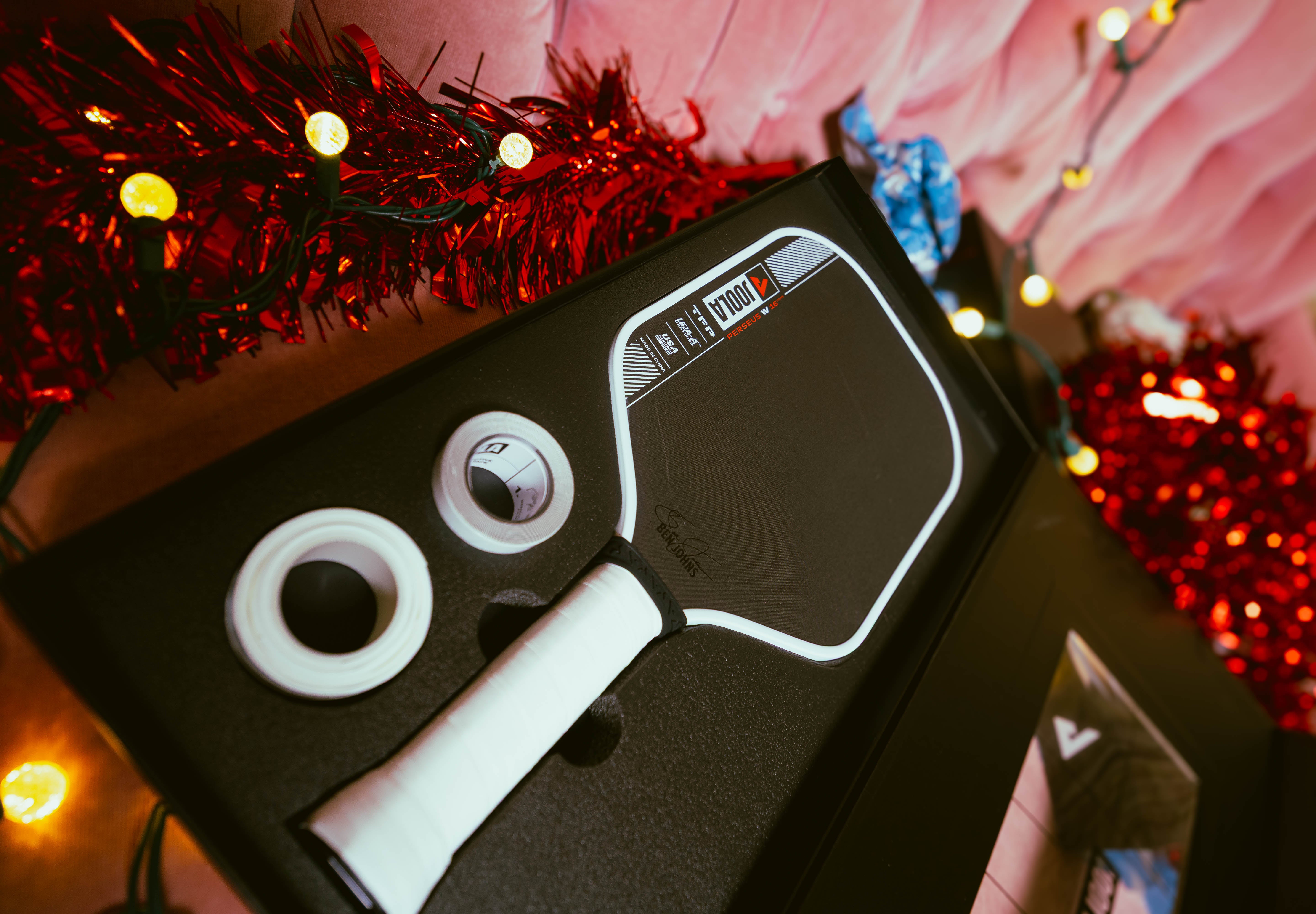 Holiday themed background, image of the JOOLA Perseus Pro IV in the box with edge guard tape and overgrip on the side.