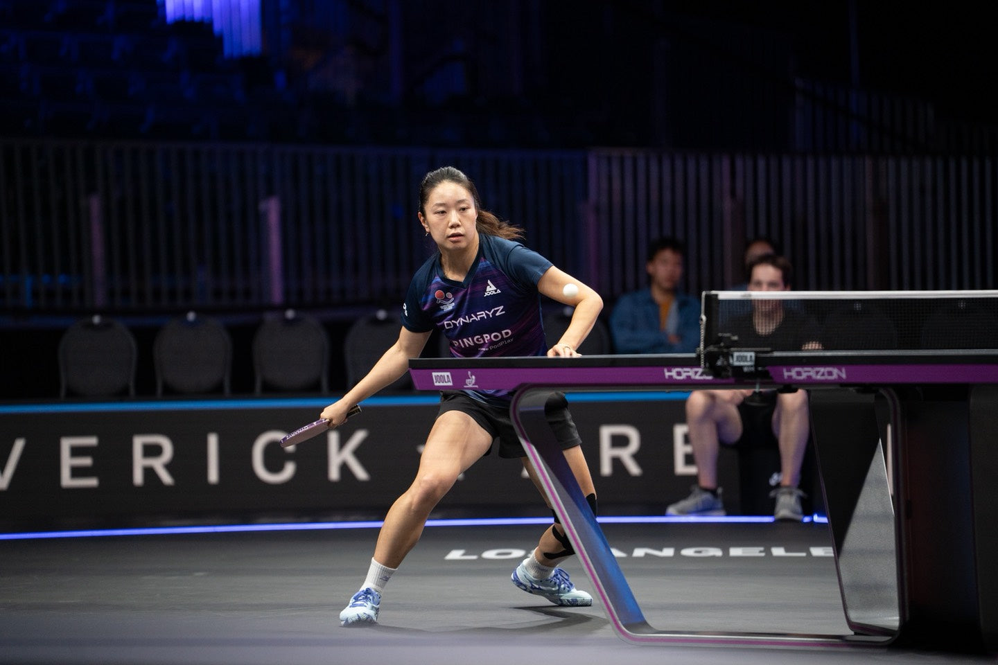 A female table tennis player in a navy and purple uniform is captured mid-swing on a professional indoor court with a JOOLA Horizon table visible in the foreground.