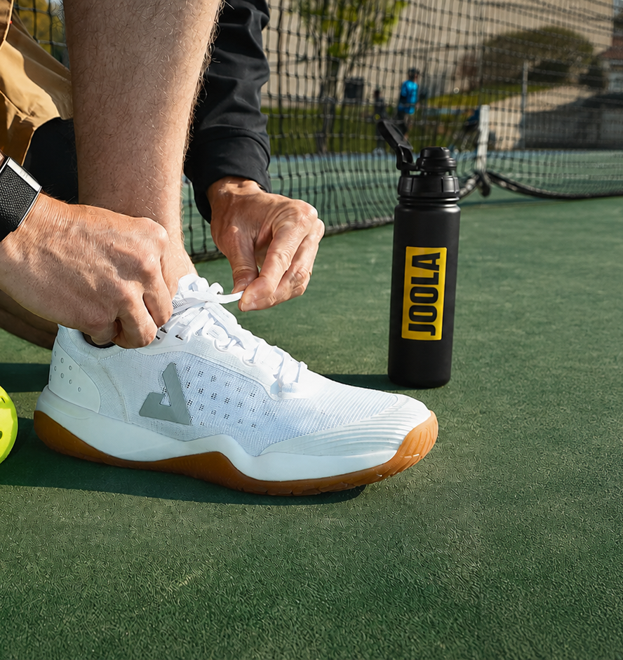 Person tying a white pickleball shoe on a pickleball court with a black water bottle labeled JOOLA in the background.