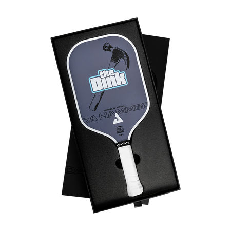 A pickleball paddle presented in an open black gift box on a white background. The paddle has a dark slate-blue face with a hammer graphic, the text 'the Dink', and 'DA HAMMER', plus a white grip.