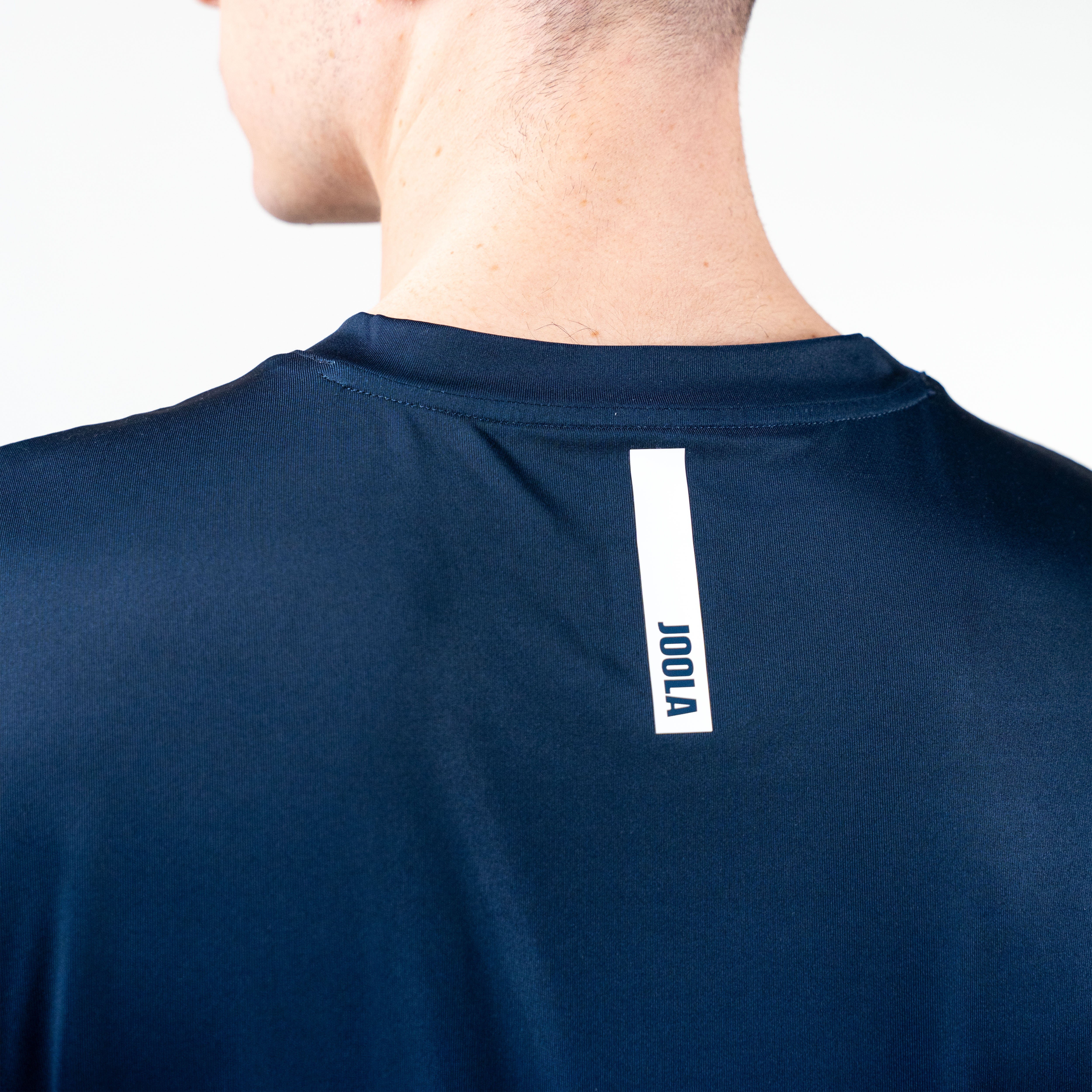 White background, close up image of the JOOLA logo on the upper back of a JOOLA Men's Competition Table Tennis shirt, blue.