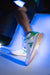 Purple and white background, image of a person wearing a pair of JOOLA TT SM4SH pickleball shoes, white and green with gum soles.