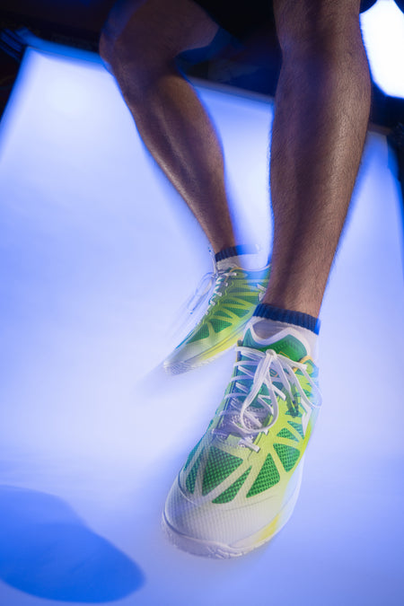 Purple-gradient colored background, image showing a model jumping up in the air wearing a pair of white, green and yellow JOOLA TT SM4SH Table Tennis shoes.