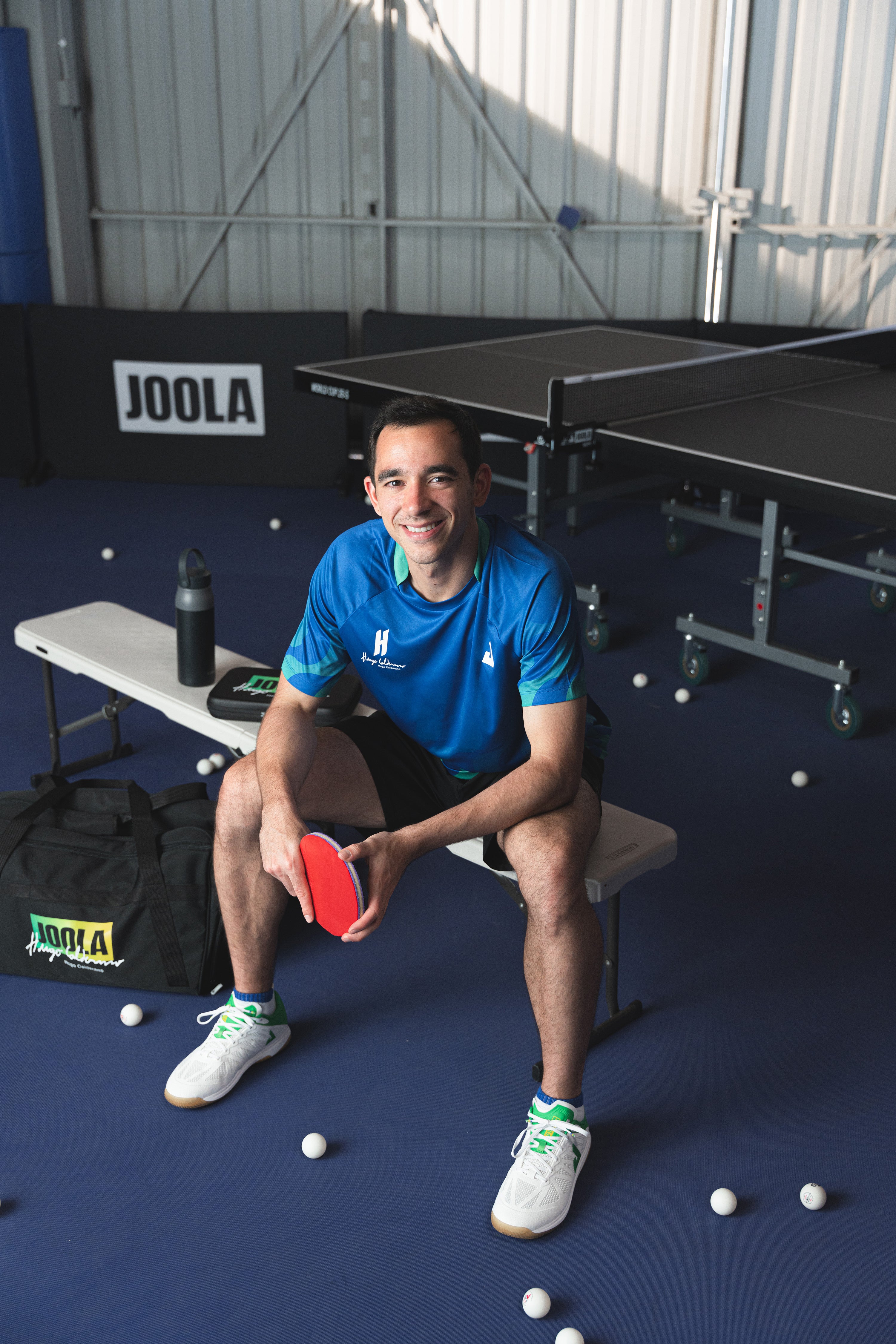 Image of JOOLA Table Tennis pro Hugo Calderano sitting on a bench at an indoor table tennis facility.