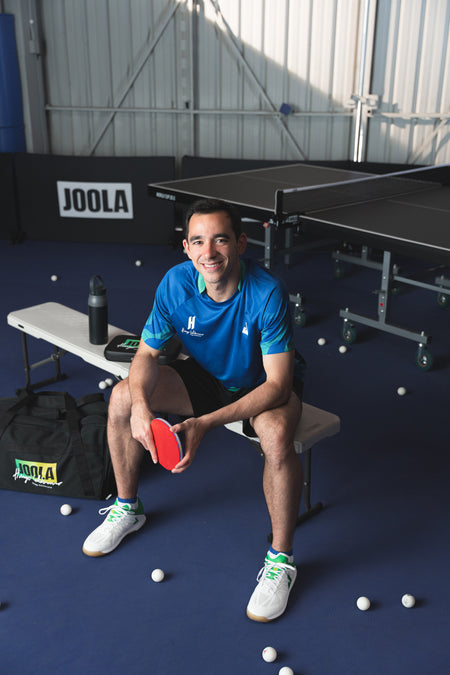 Image of JOOLA Table Tennis pro Hugo Calderano sitting on a bench at an indoor table tennis facility.