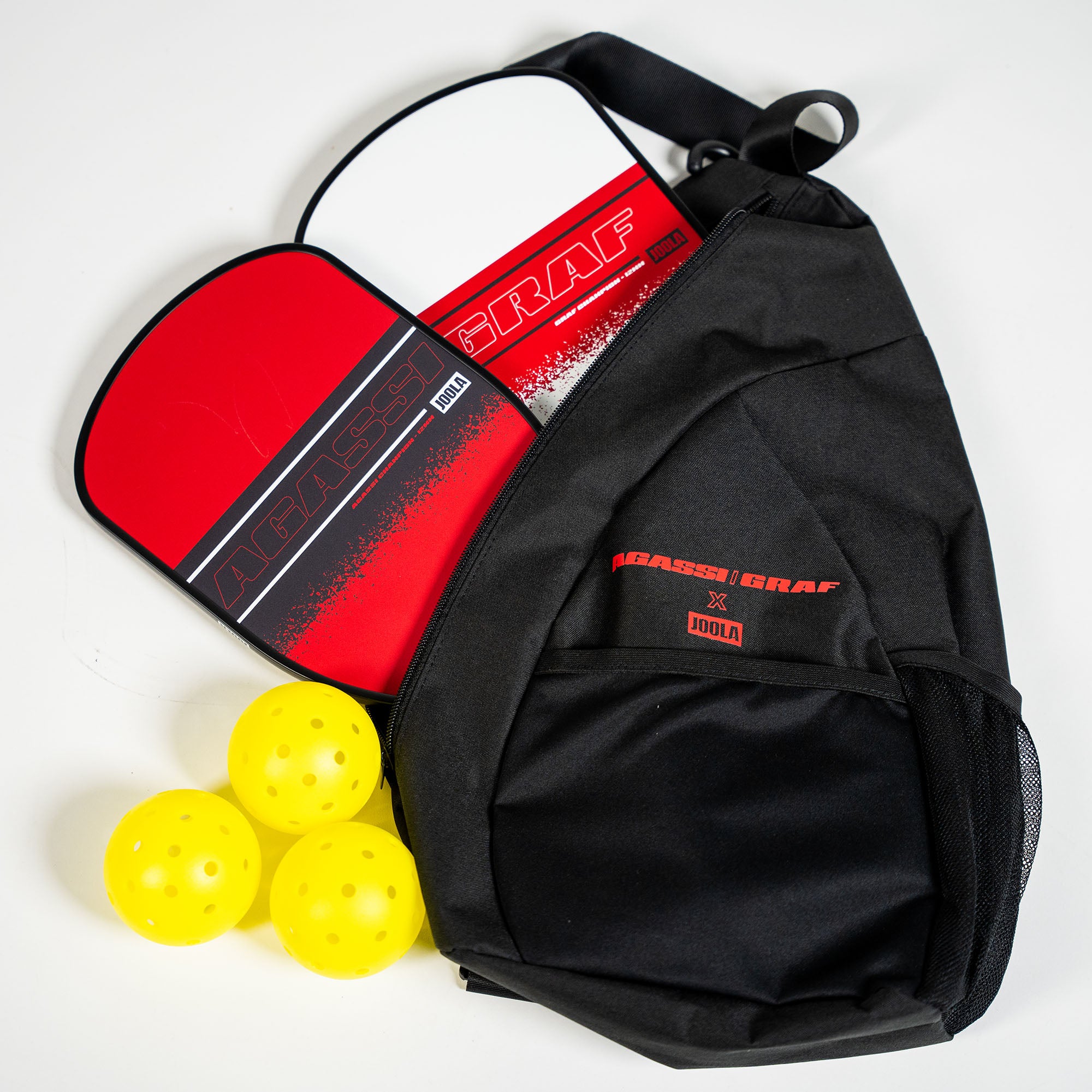 White background, product image showing the Agassi x JOOLA Champion pickleball paddle set. One Agassi Champion Paddle, One Graf Champion Paddle, three Heleus pickleballs and a Agassi/Graf x JOOLA sling bag.