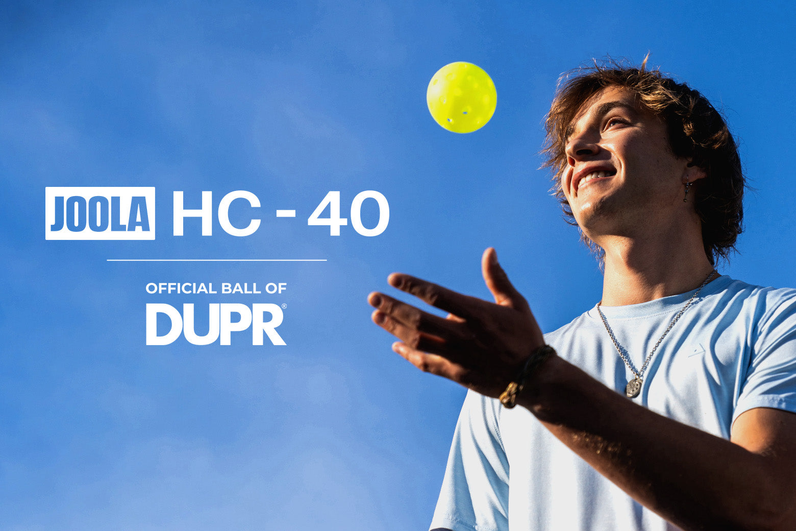 A man in a light blue t-shirt looking up at a yellow Joola HC-40 ball against a bright blue sky, with text reading 'JOOLA HC - 40' and 'OFFICIAL BALL OF DUPR'.