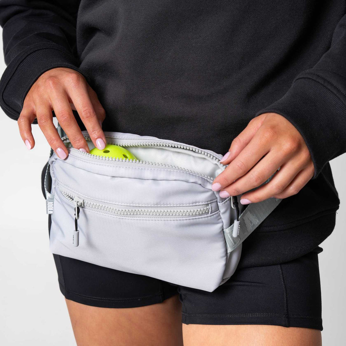 Close up image of a person putting a pickleball in to the pockey of a JOOLA Belt Bag, Gray.