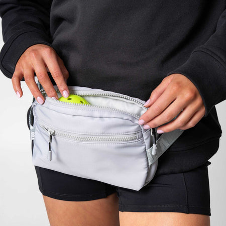 Close up image of a person putting a pickleball in to the pockey of a JOOLA Belt Bag, Gray.