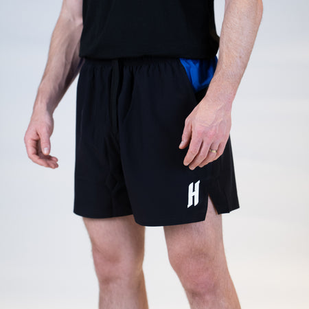 White background, product image of the JOOLA Hugo Calderano shorts, black and blue with a 5-inch inseam. The image shoes the stylized H of the Hugo Calderano brand.