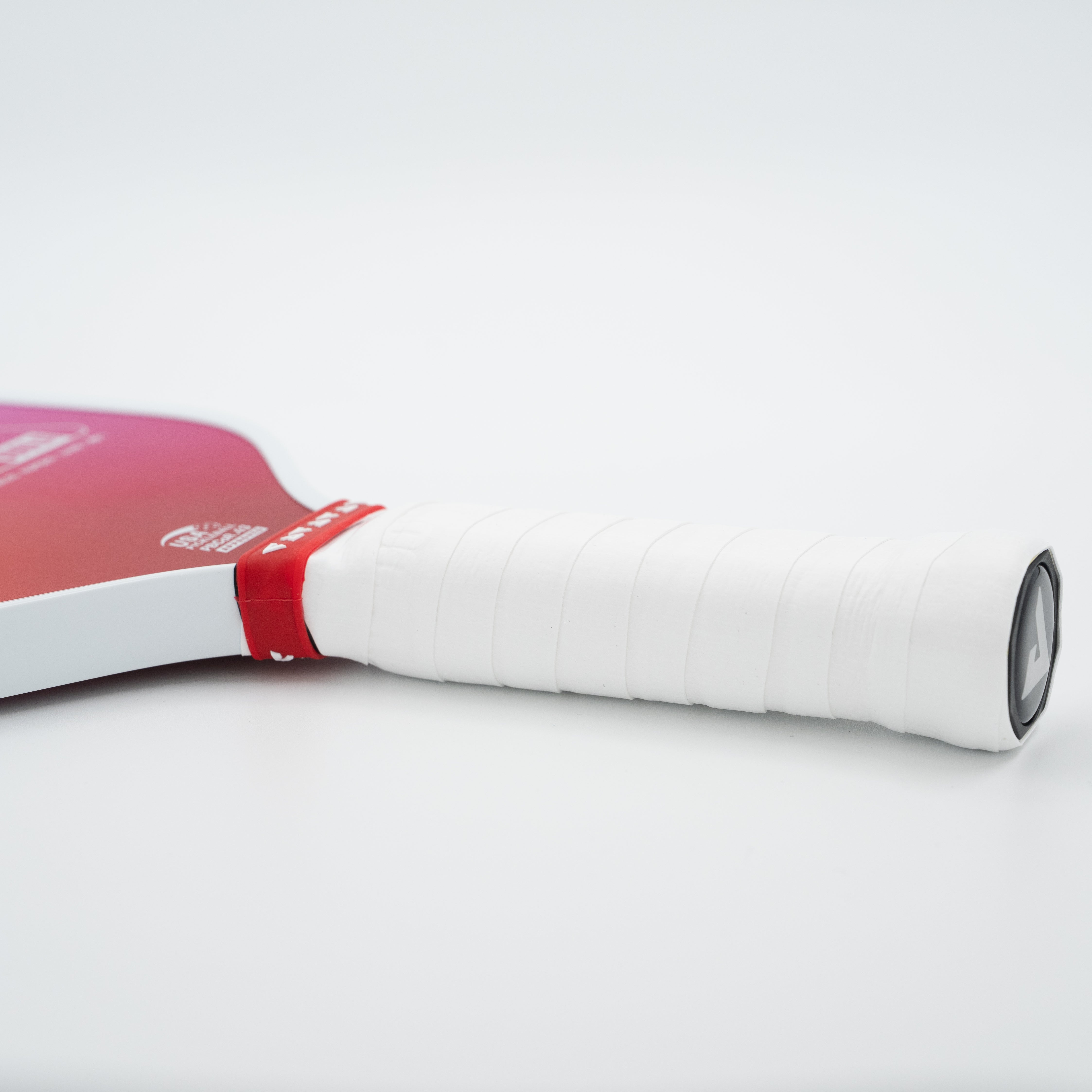 A close-up of a JOOLA pickleball paddle handle wrapped in white grip tape with a red band, showing part of the red and white blade against a clean white background.