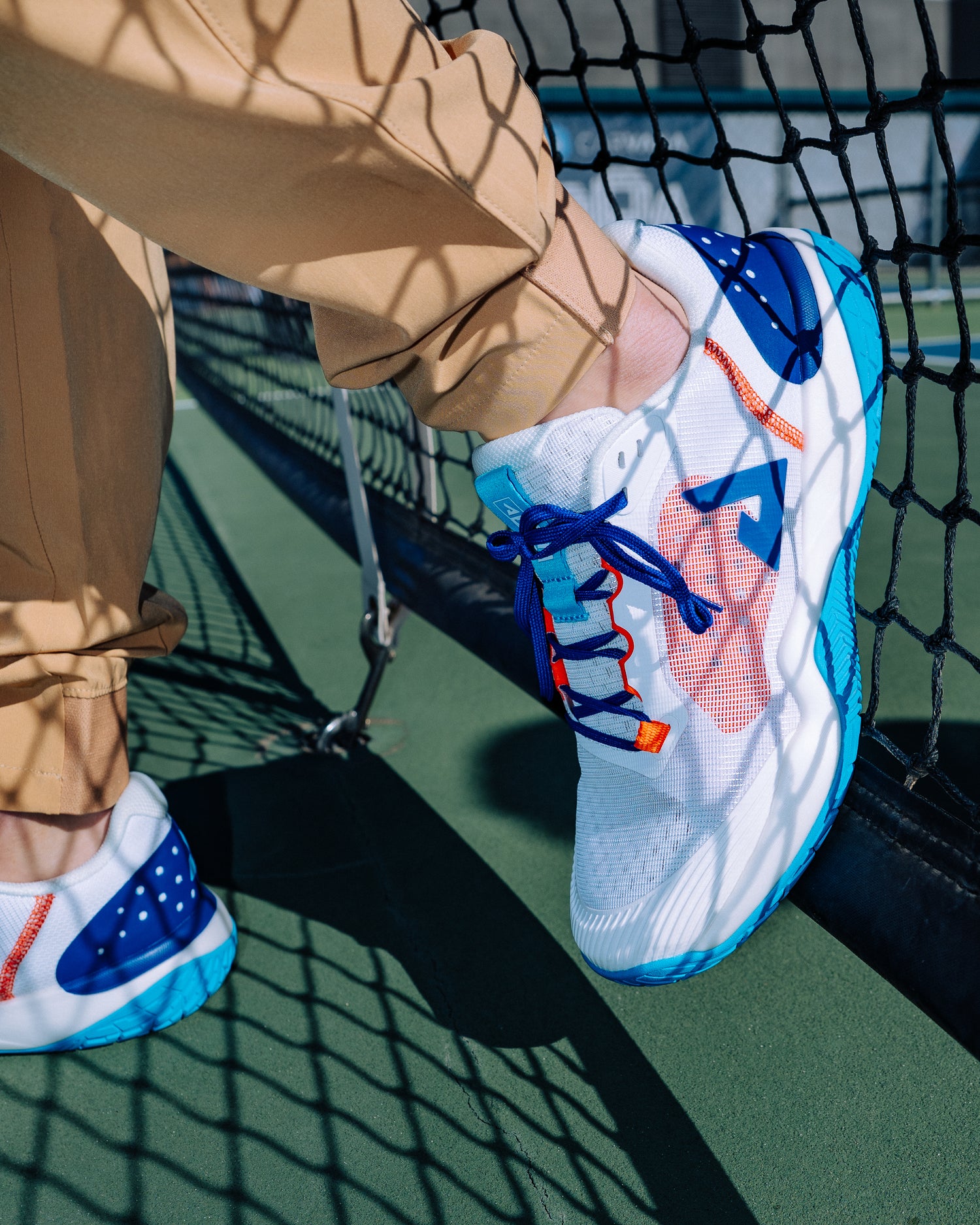 Closer view of the JOOLA R4LLy Blue shoes against a background of a pickleball net.