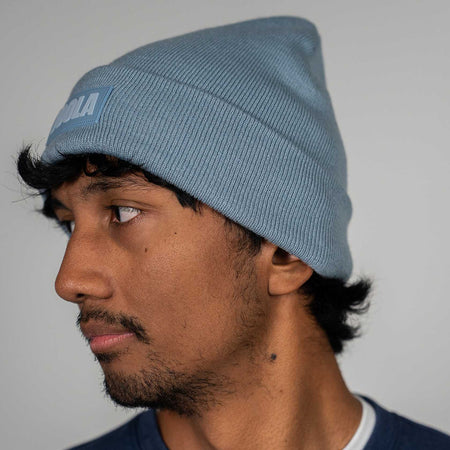 Person wearing a dusty blue winter beanie with a brand logo on a plain background