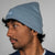 Person wearing a dusty blue winter beanie with a brand logo on a plain background
