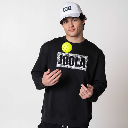 Gray background, image of a male model tossing a pickleball up in the air wearing a JOOLA Fleece, black.