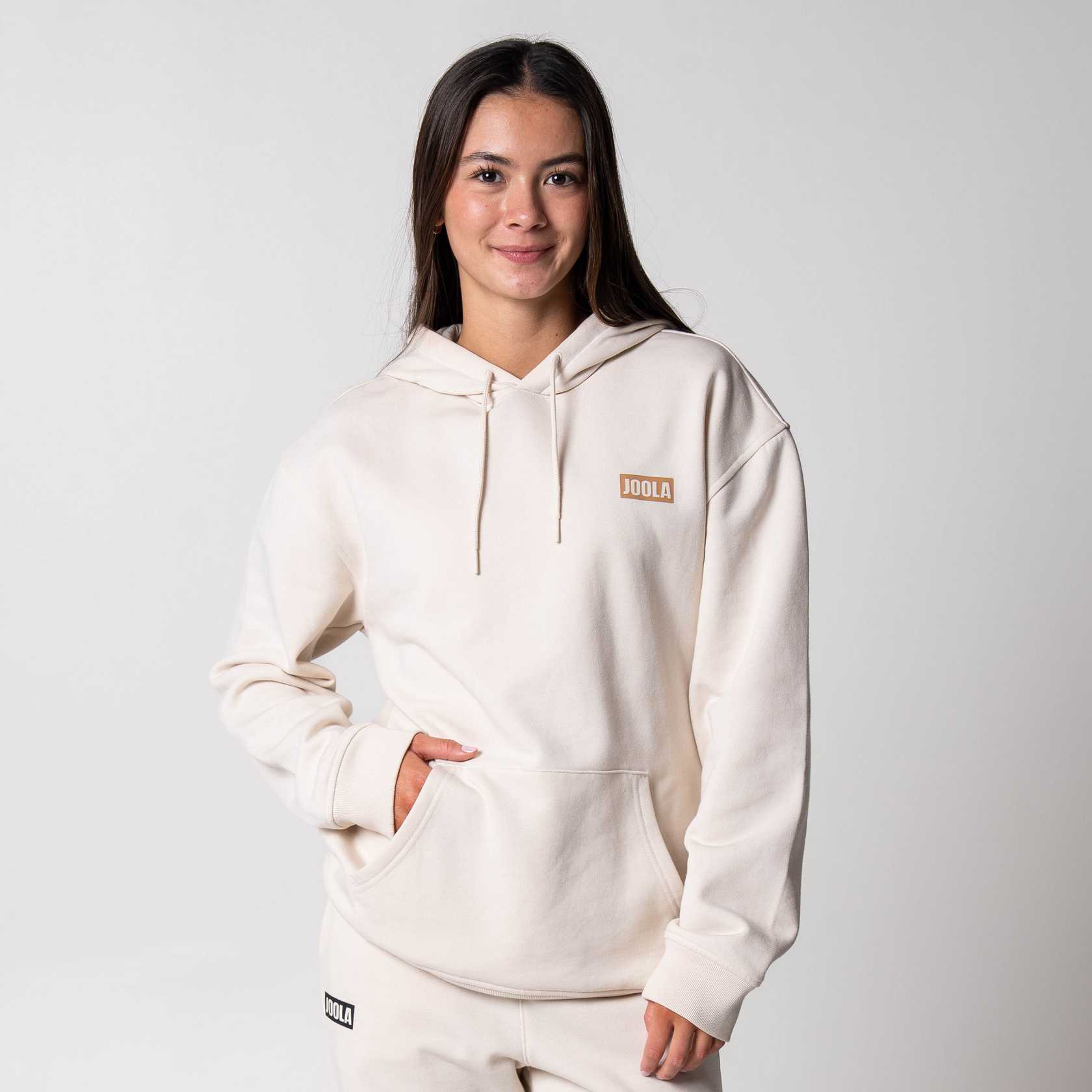 Gray background, image of a female model wearing a Sandstone colored JOOLA Fleece Hoodie.