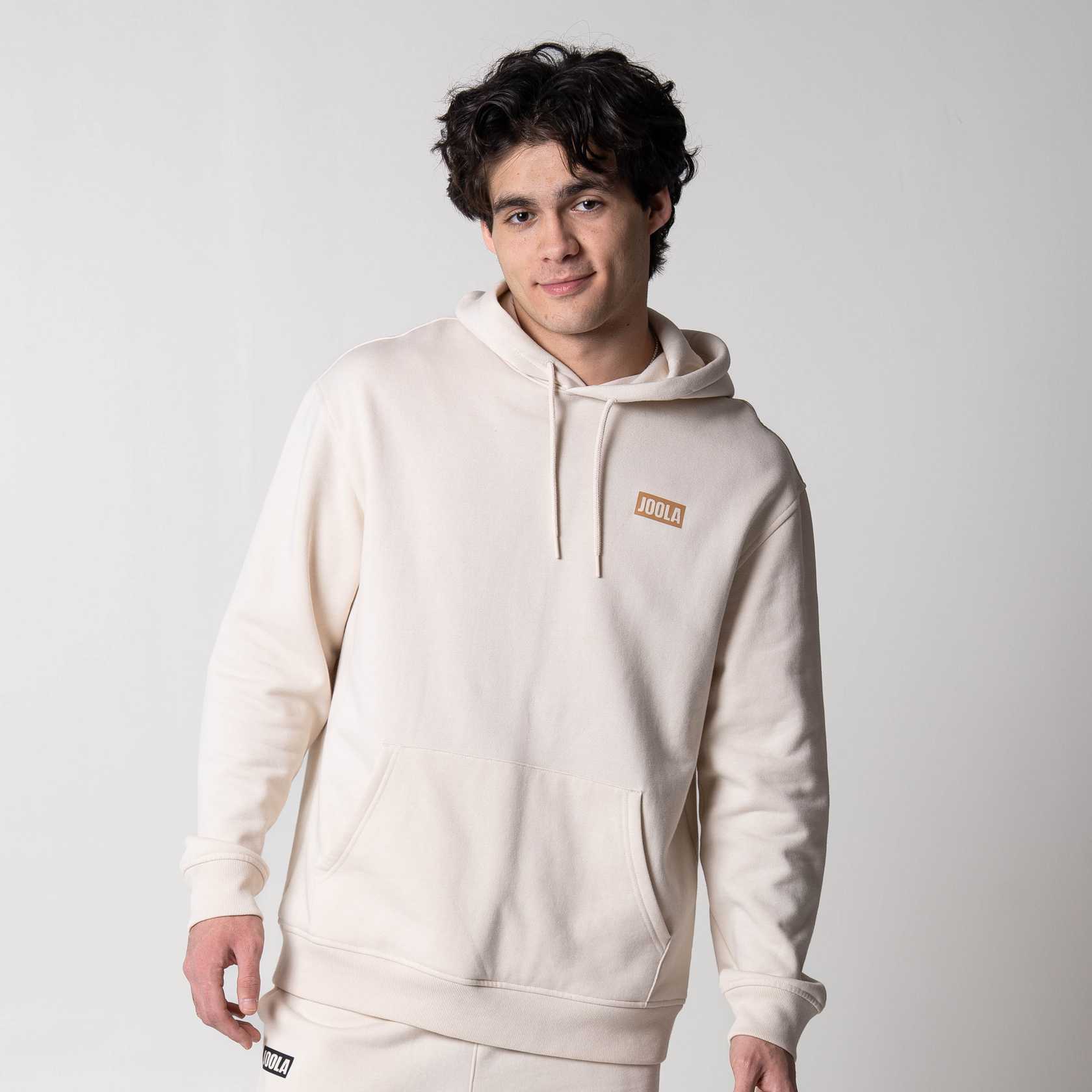 Gray background, image of a male model wearing the JOOLA Fleece Hoodie, Sandstone Colored.