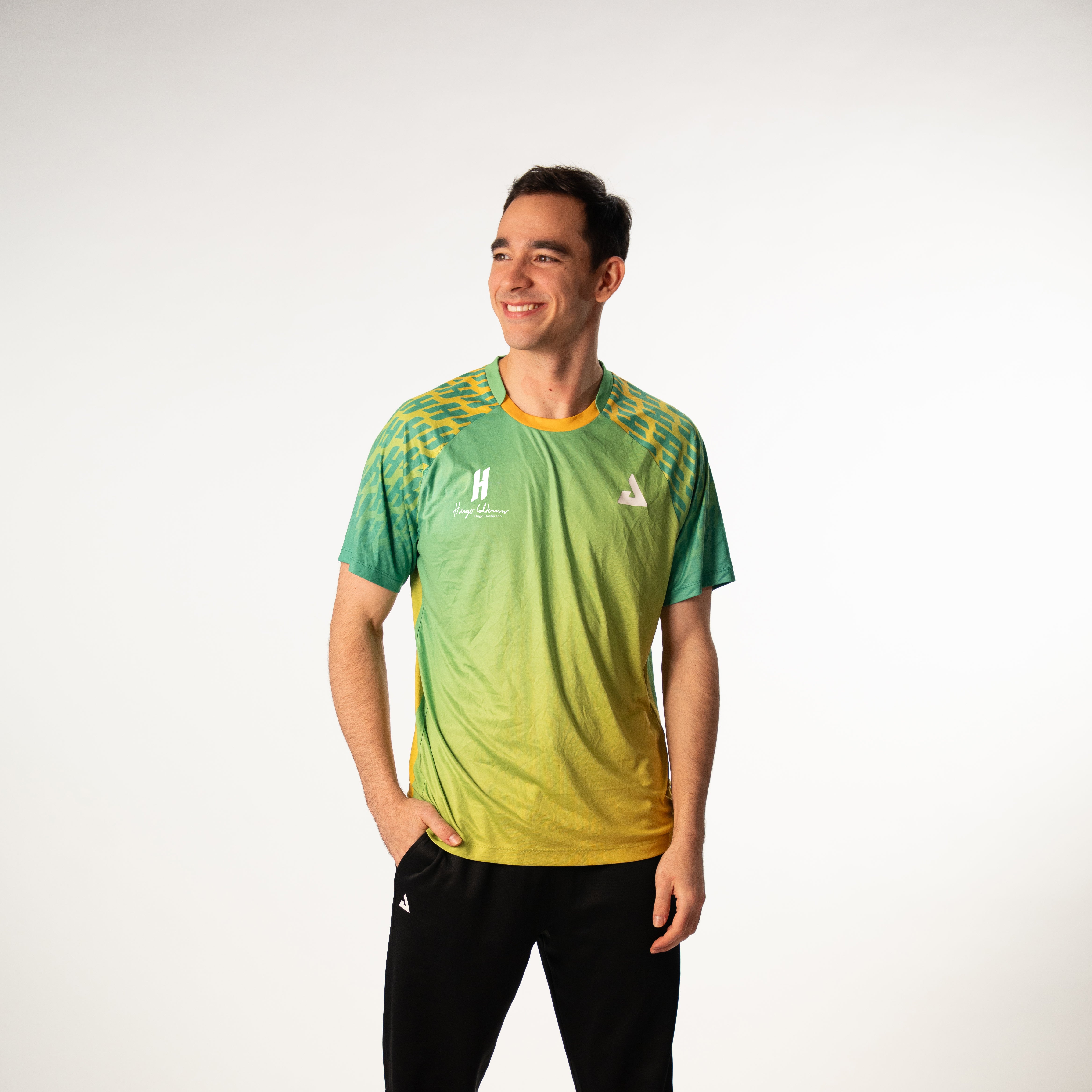 White background, image of Hugo Calderano wearing a Hugo Calderano Gradient Monogram Raglan Short Sleeve shirt. Jungle Green colorway.
