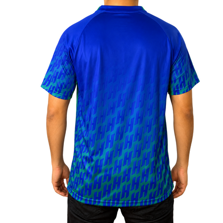 White background, image showing the back of a male model wearing a Hugo Calderano Monogram Crossover Raglan Short Sleeve shirt, azul colorway.