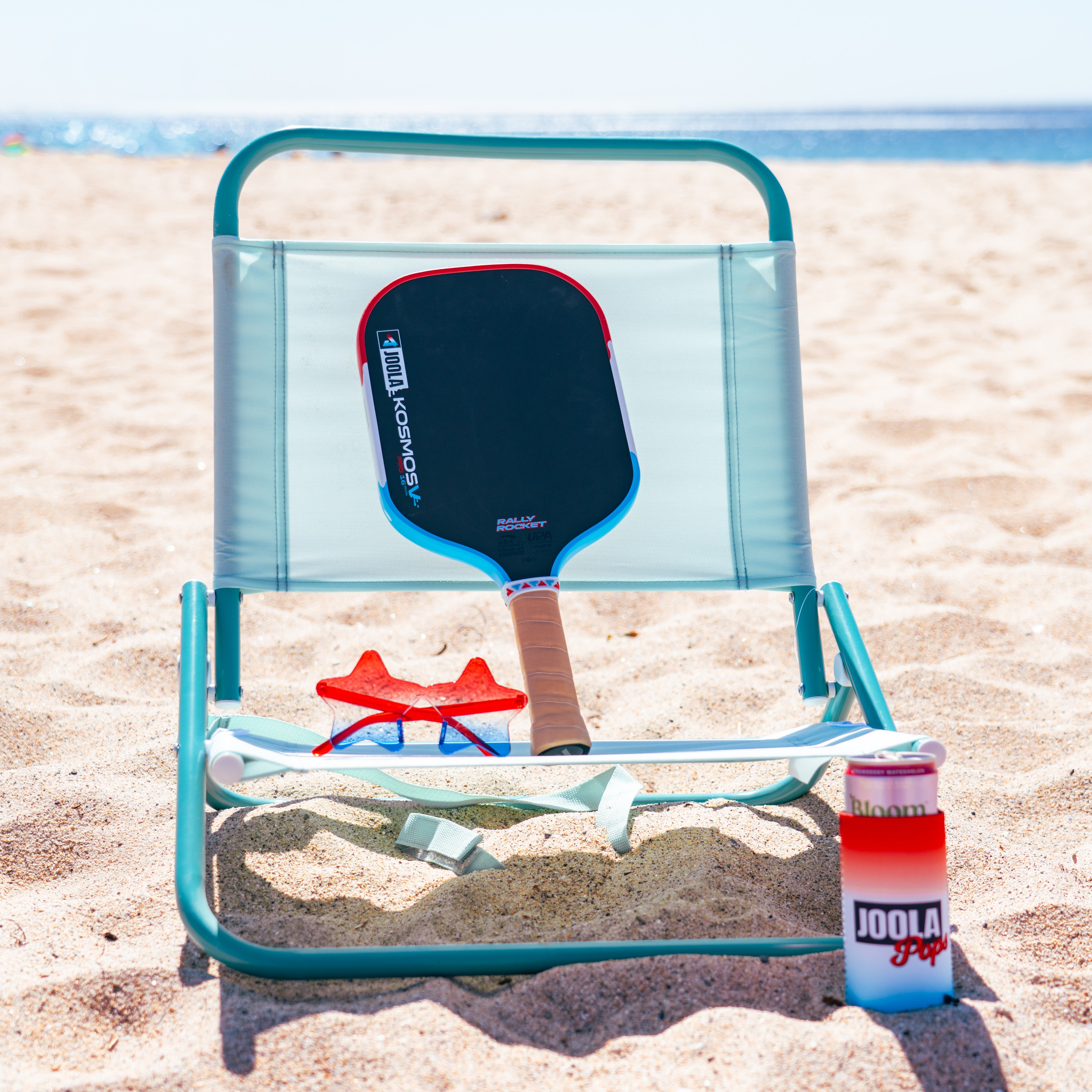 Blue folding chair with a JOOLA Kosmos Rally Rocket pickleball paddle, JOOLA pops coozie on a beach. 