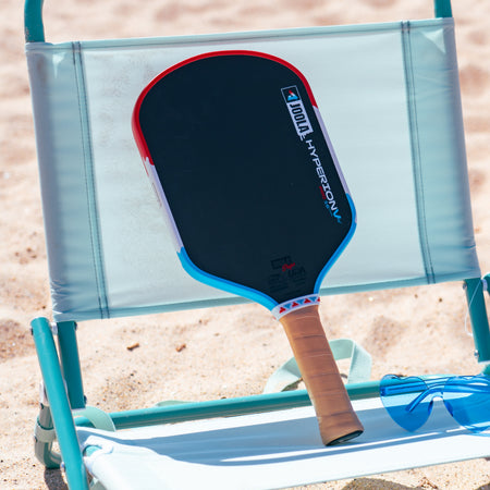 JOOLA Rally Rocket Hyperion Pro V pickleball paddle sitting on a beach chair on the beach with a pair of blue heart sunglasses next to the paddle.