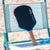 JOOLA Rally Rocket Hyperion Pro V pickleball paddle sitting on a beach chair on the beach with a pair of blue heart sunglasses next to the paddle.