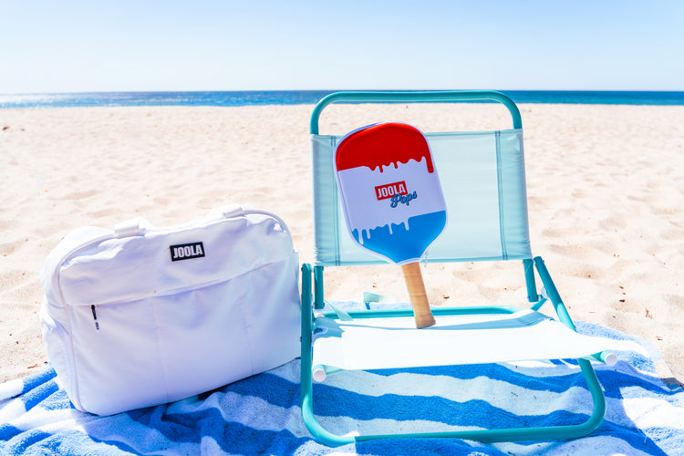 Beach chair with a blue and red paddle  cover over a JOOLA R4LLy Rocket pickleball paddle and white bag on a sandy beach.