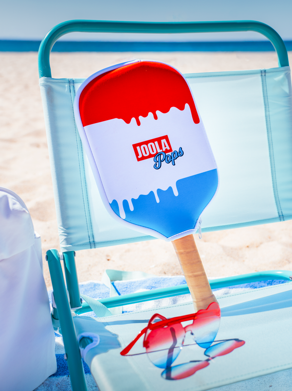 Red and blue JOOLA Pops paddle on a beach chair with sunglasses on a sand background.