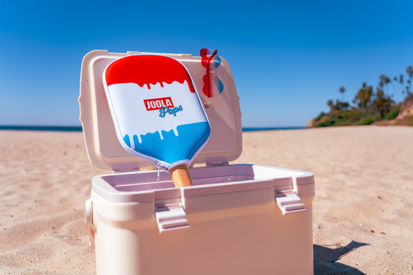 JOOLA Pops paddle cover on a JOOLA Rally Rocket pickleball paddle sitting in a cooler on a beach.