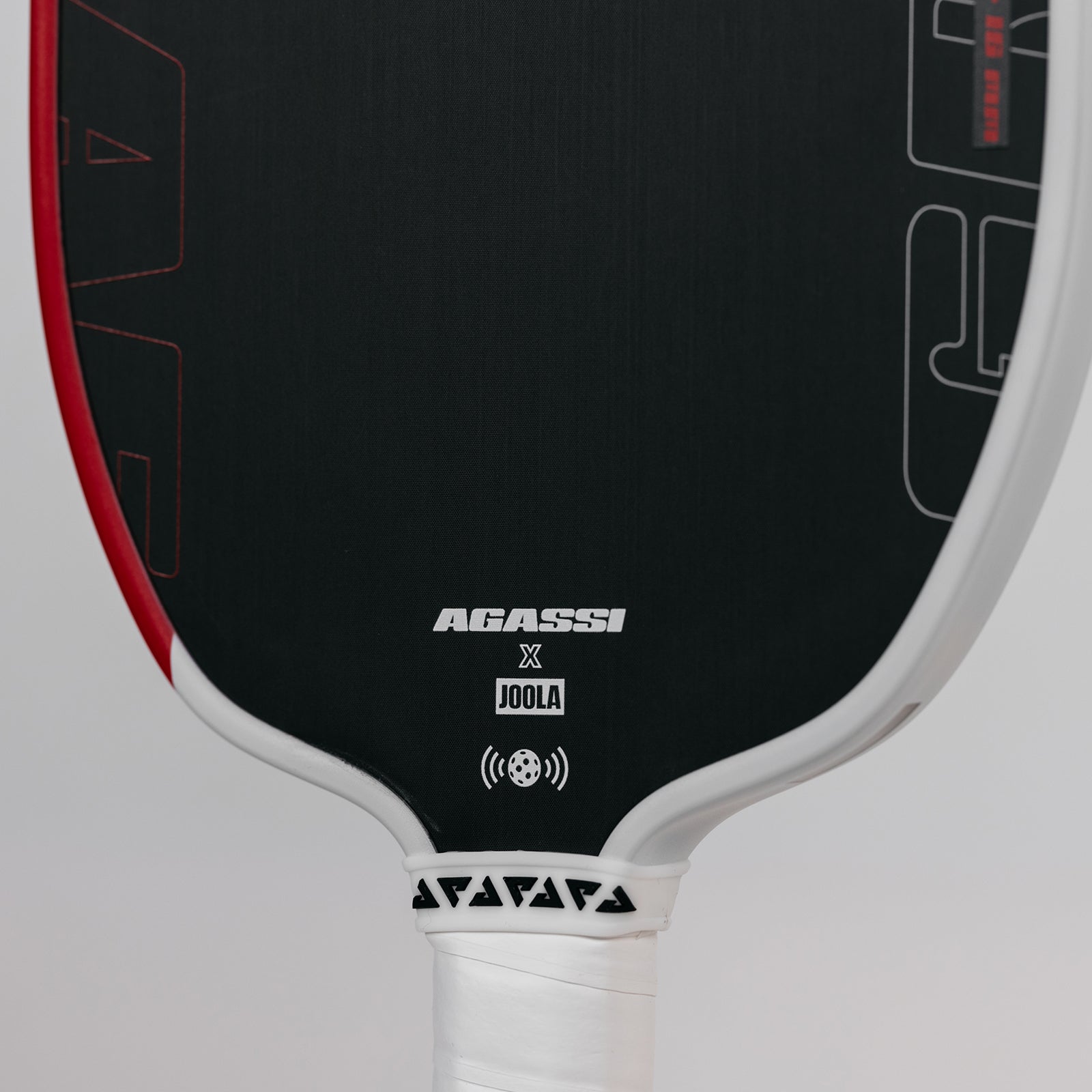 Close up image of the Agassi x JOOLA near the base of the Agassi x JOOLA Graf pickleball paddle. Black surface, white handle, red and white edge guard.