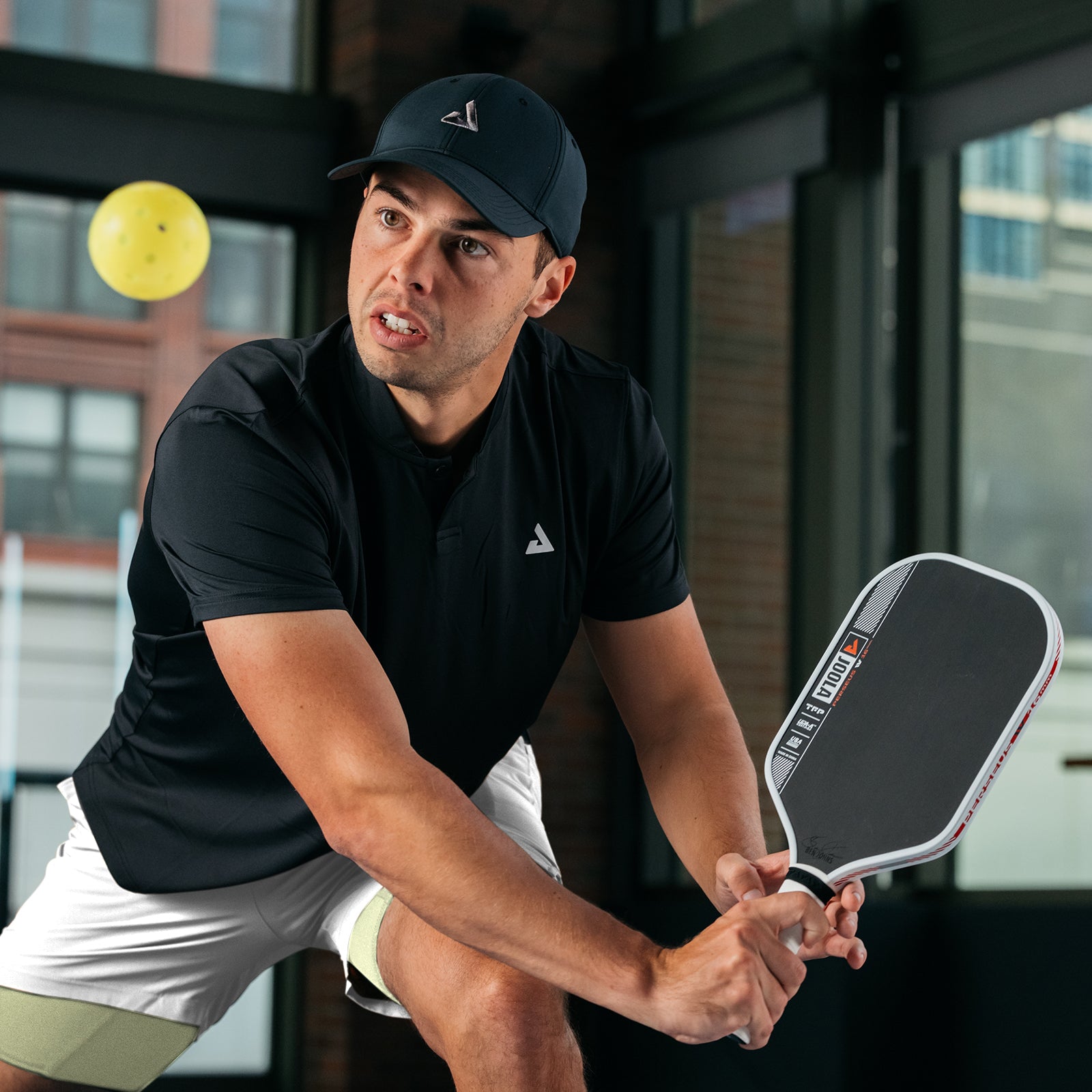 Johns Perseus Pro IV pickleball paddle. He appears in JOOLA apparel on an indoor pickleball court in front of an apartment style floor to ceiling window.