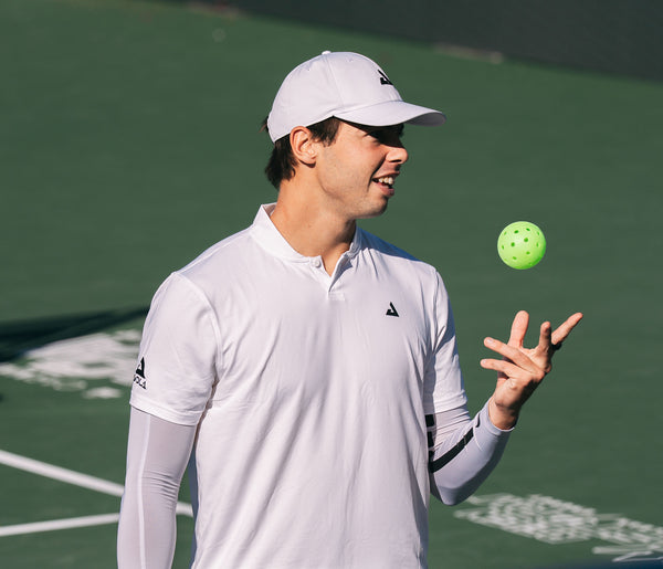 Image of JOOLA pro Ben Johns tossing a pickleball in the air and wearing a white JOOLA polo and white JOOLA hat.