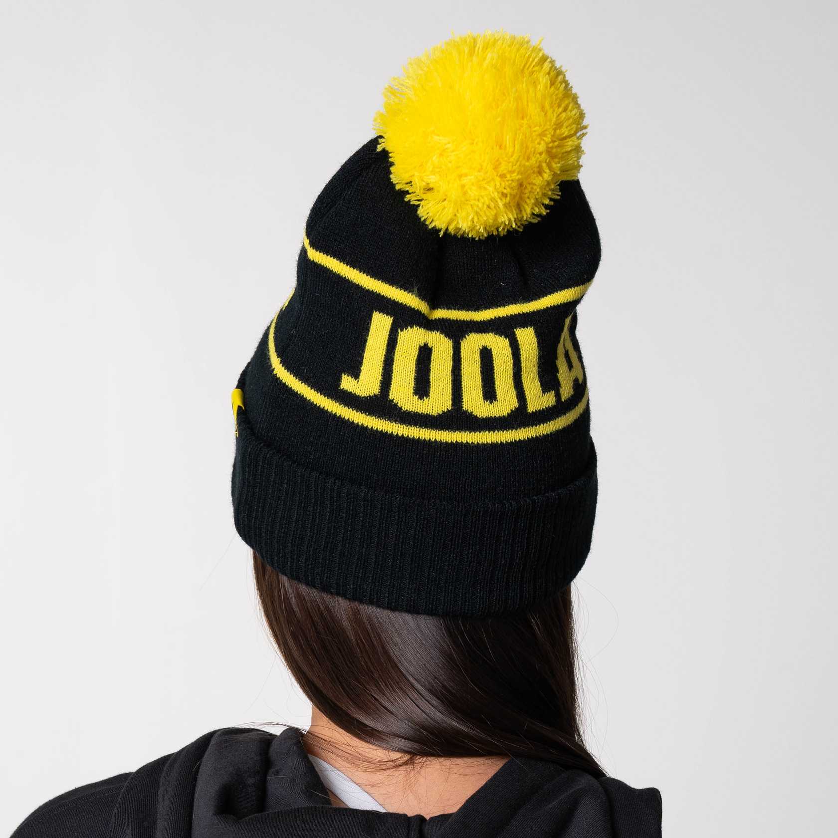 Gray background, image of a model wearing a black and yellow JOOLA Pom Pom Beanie.