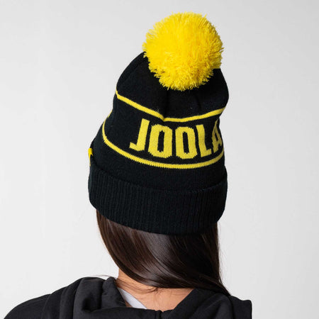 Gray background, image of a model wearing a black and yellow JOOLA Pom Pom Beanie.