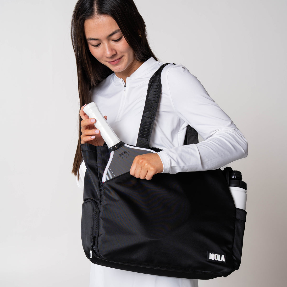 Gray background, image of a femal model putting a JOOLA pickleball paddle in her JOOLA overnight bag.
