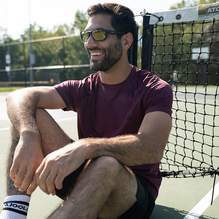 Male model sitting on the pickleball court with his back resting on the net, male model is wearing JOOLA short sleeve shirt, woven shorts, tube socks and JOOLA RJX eyewear.