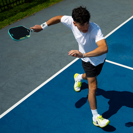 A man playing pickleball on a blue court, holding a JOOLA Power FX Daydream colorway. He is wearing a white t-shirt, black shorts, white socks, and white sneakers with neon yellow accents.
