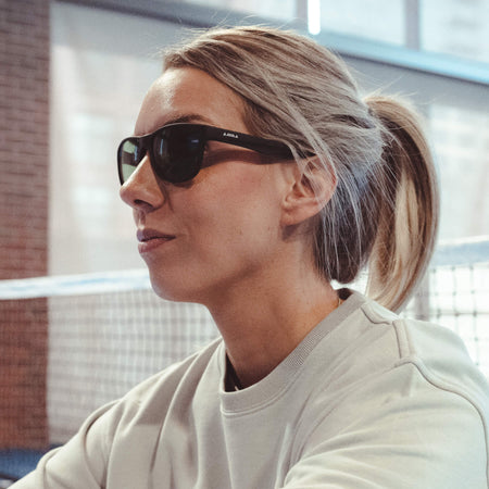 Close up image of a female model wearing JOOLA indoor eyewear sitting on the pickleball court posing.